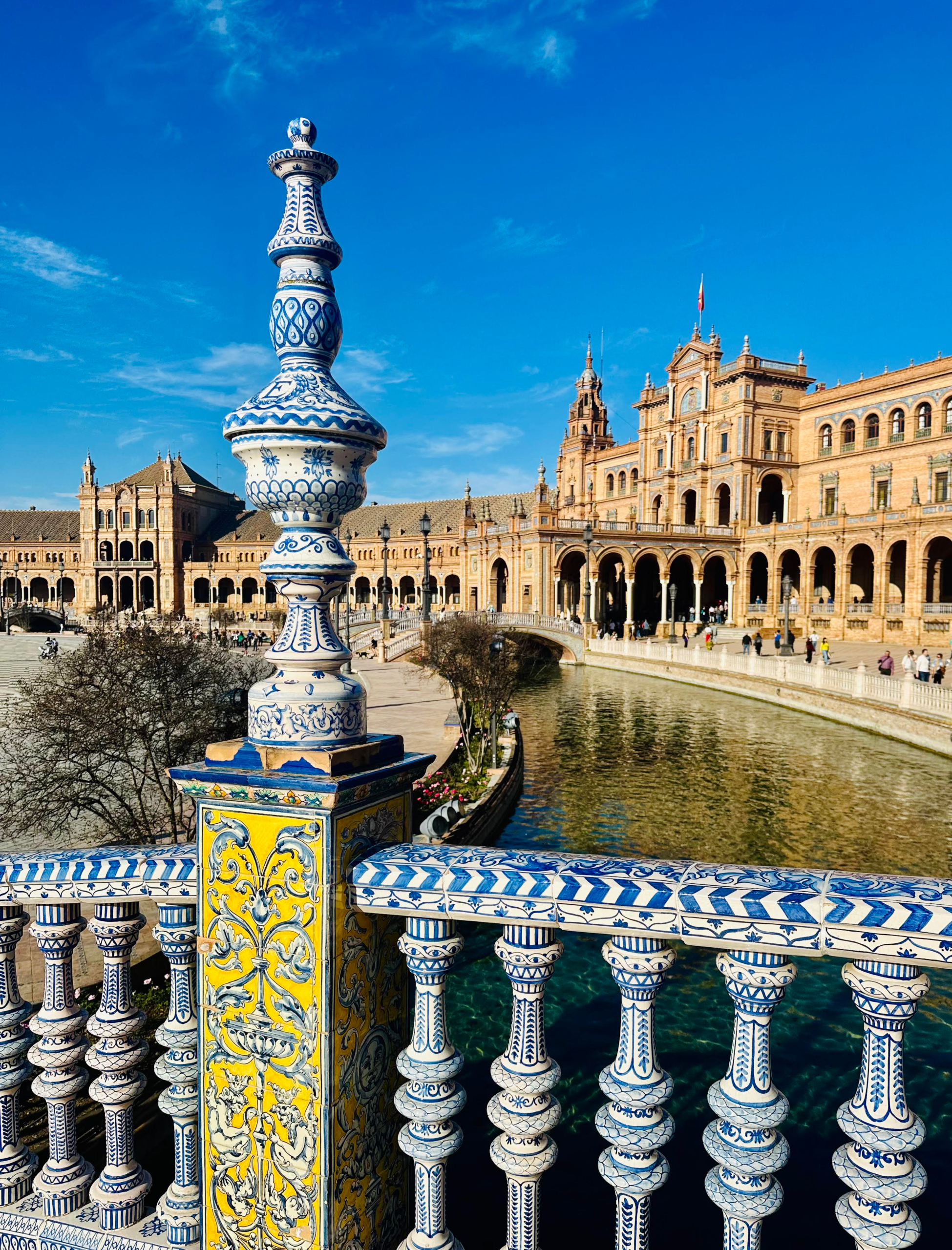 Wide view of Seville’s Plaza de España with its grand curved building, ornate bridges over the canal, under a sunny sky.